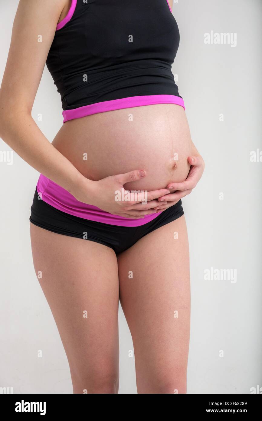 Young adult pregnant girl in sportswear. Sport and pregnancy concept. Yoga and fitness in anticipation of childbirth. White backdrop. Stock Photo