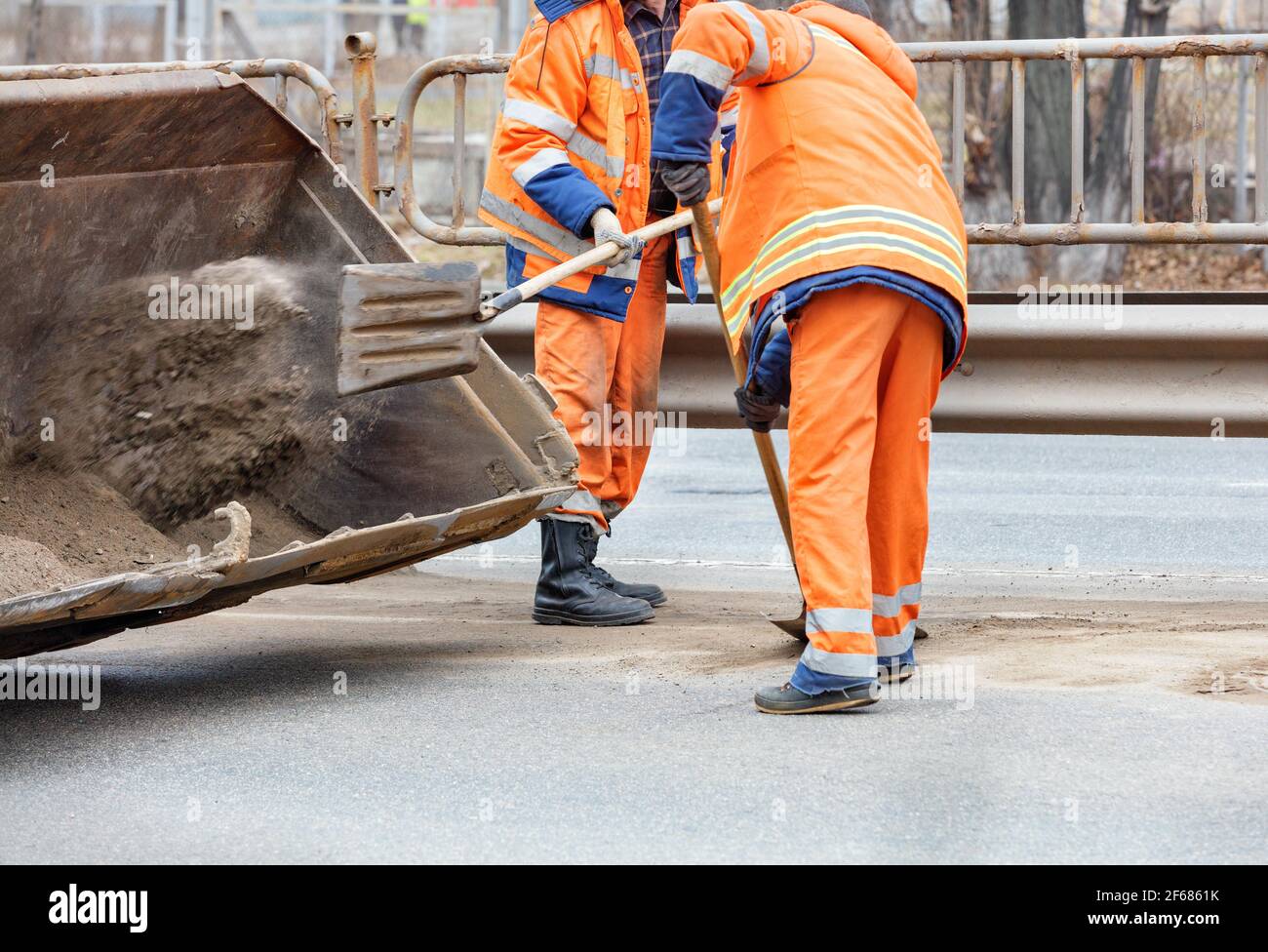 Road workers scrape off accumulated sand between lanes of the road with shovels and load it into a metal grader bucket. Stock Photo