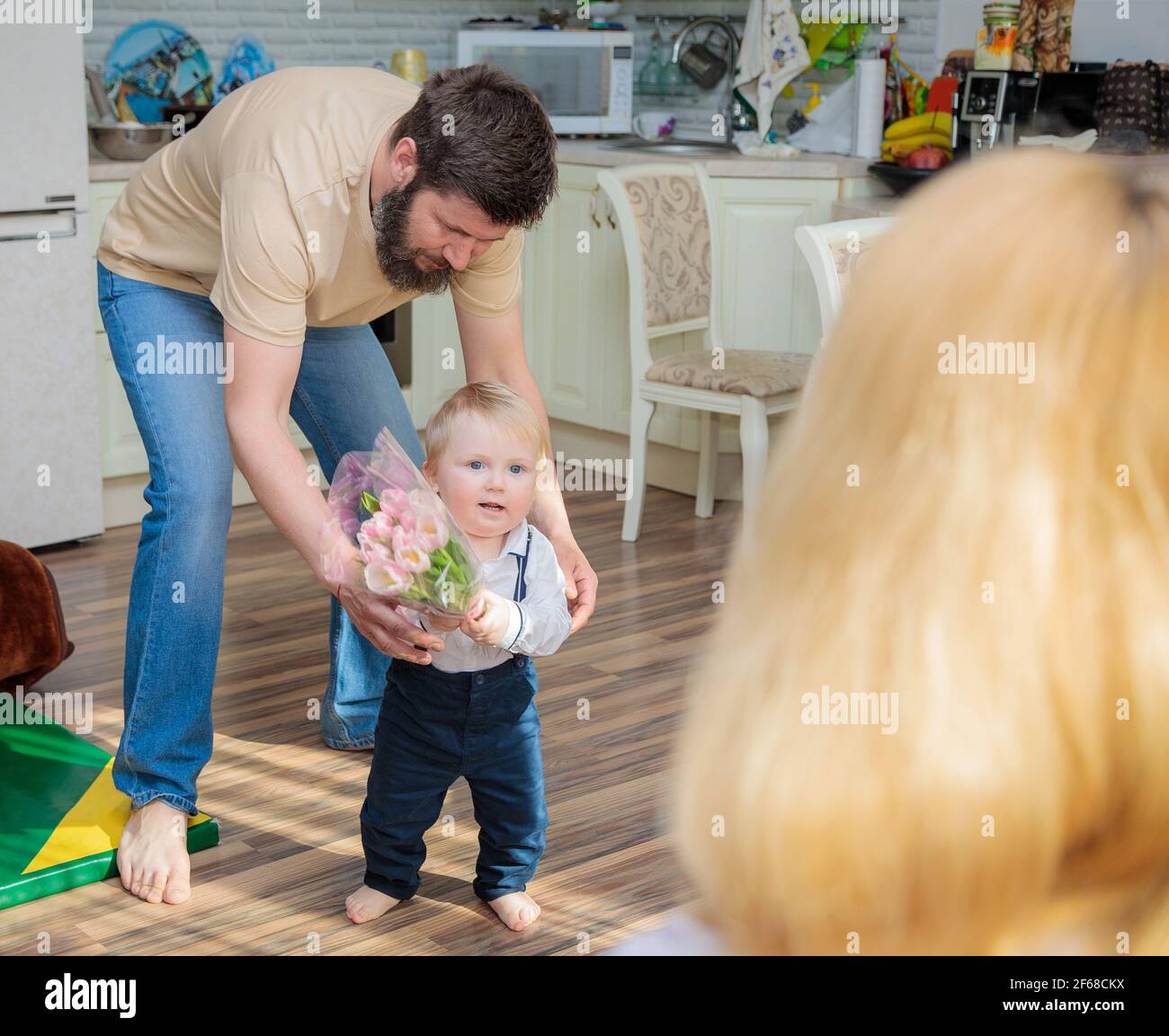 Dad with baby, son congratulate mom on the holiday. The kid brings his mother a bouquet of flowers and a gift. Stock Photo