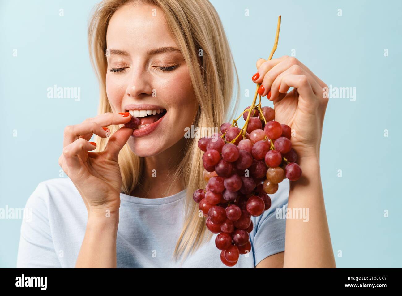 Happy charming blonde girl eating grapes on camera isolated over blue