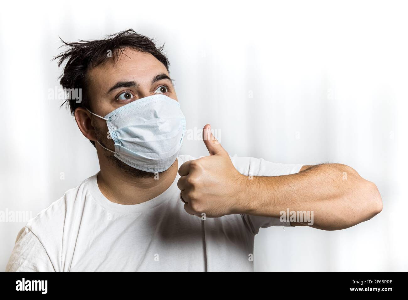 An unshaven man in a medical mask and a white T-shirt with tousled hair looks to the right and up and shows a gesture - thumbs up. On white background. Stock Photo