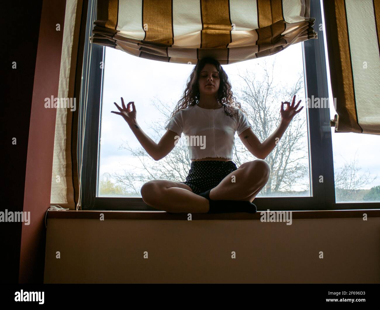 young woman sitting on a window looking out into backlight doing yoga Stock Photo
