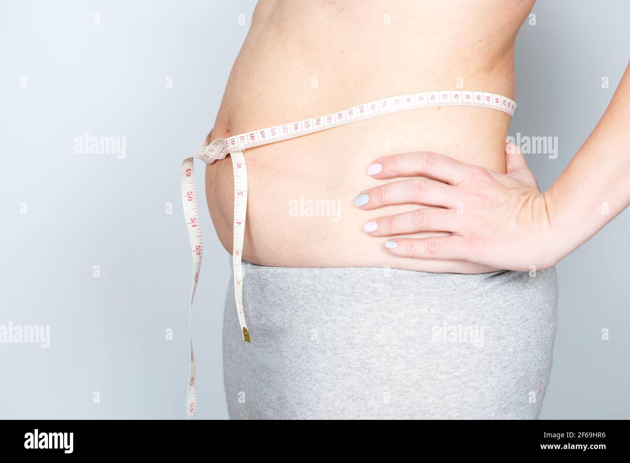 Overweight or fat adult woman is measureing around the body on the midriff on a gray background. Healthcare, medicine concept. Stock Photo
