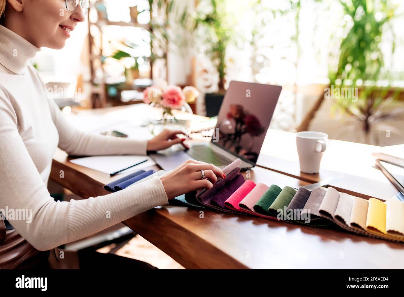 An interior designer is working on a new project. Choosing color from fabric color palette for new furniture. Oak wooden table. Cup of coffee, Flowers Stock Photo