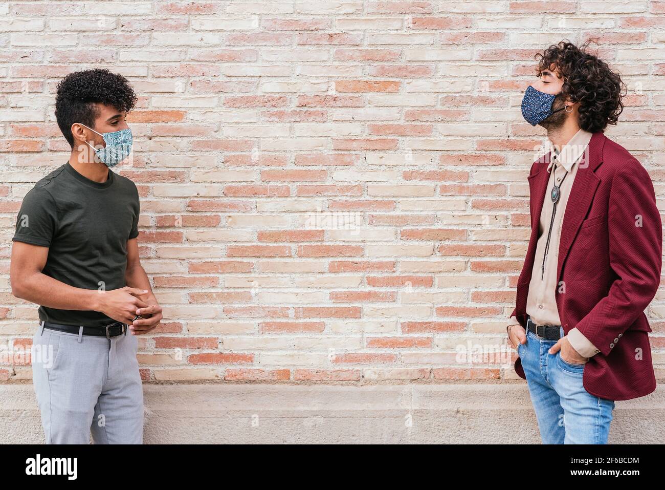 latin and caucasian man standing near a brick wall and keeping security distance. They wear casual clothing and protective face masks. Stock Photo