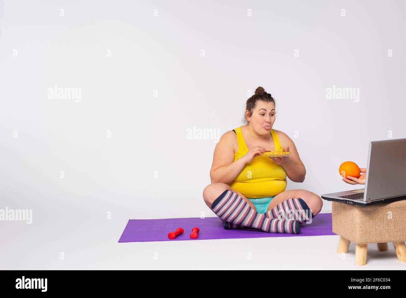 Young fat caucasian woman bending over a sports rug pointing her tongue at the laptop with hand holding orange fruit, eating sweets. A chubby girl Stock Photo