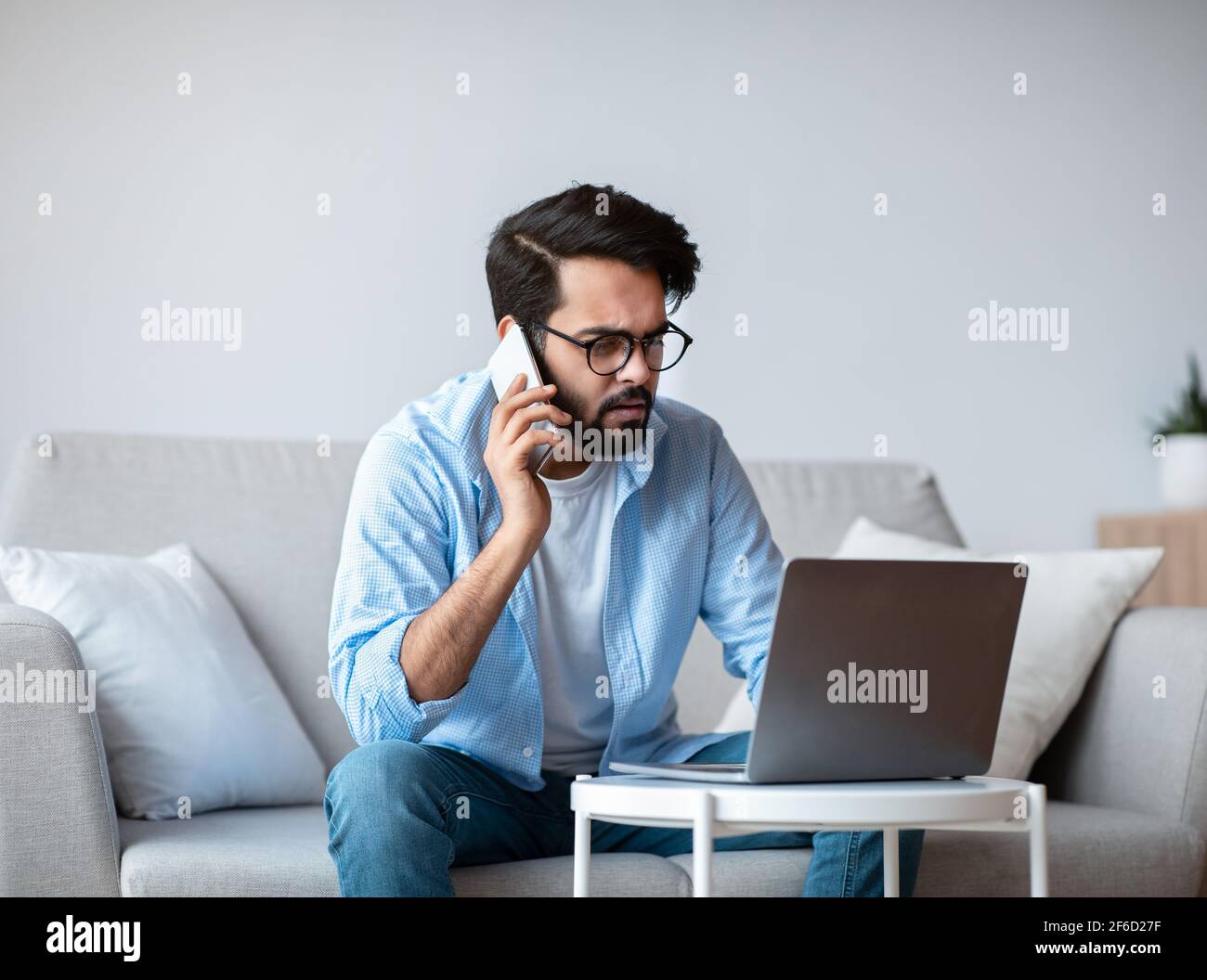Busy Eastern Freelancer Guy Talking On Phone And Using Laptop At Home Stock Photo