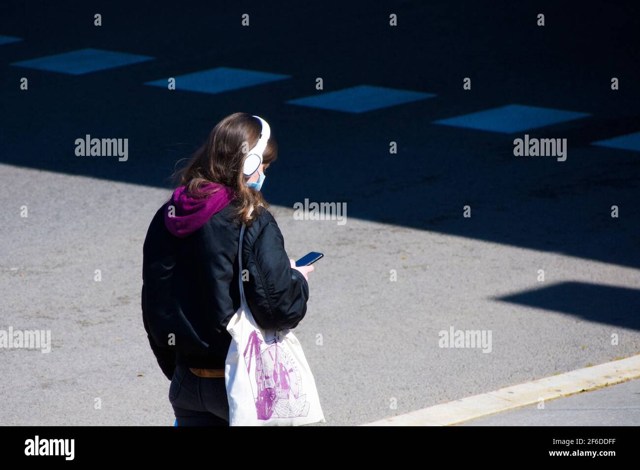 young,woman,mask,headphones,smartphone,street, walking,sunny, light, city,urban,scene,bag,jacket,lifestyle,close-up,listening,music,protection,covid Stock Photo