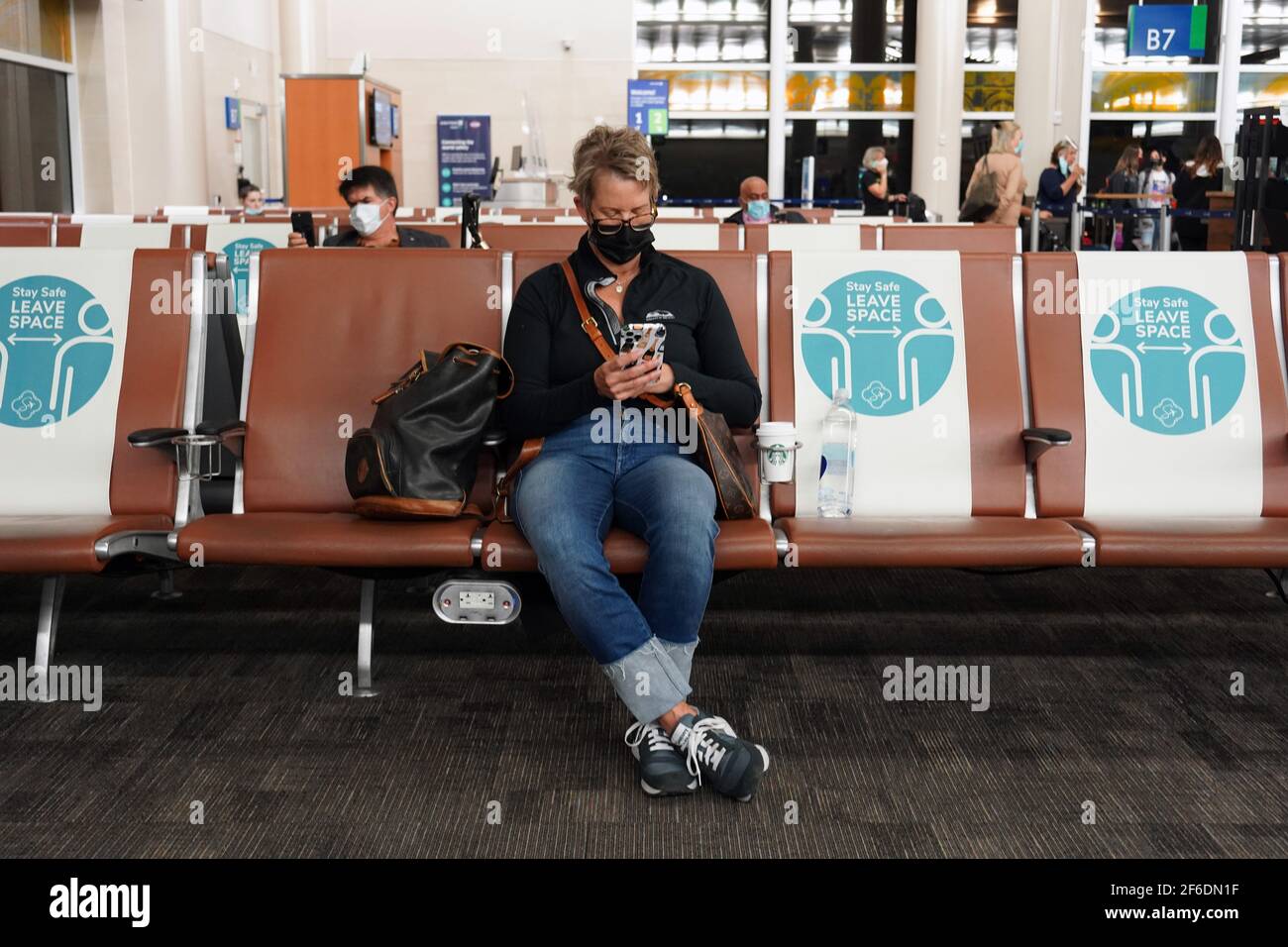 People with face masks sit between physical and social distancing marker signs with the words 'Stay Safe. Leave Space'  on seats in Terminal B of the Stock Photo