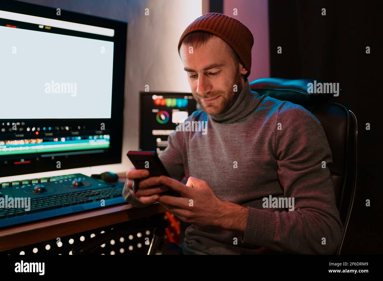Attractive Male Video Editor Works with Footage or Video on His Personal Computer and having a break communicating on his smartphone. He Works in Creative Office Studio or home. Neon lights Stock Photo