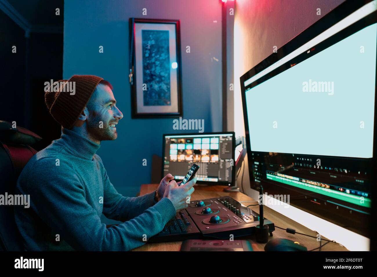 Attractive Male Video Editor Works with Footage or Video on His Personal Computer and having a break communicating on his smartphone. He Works in Creative Office Studio or home. Neon lights Stock Photo