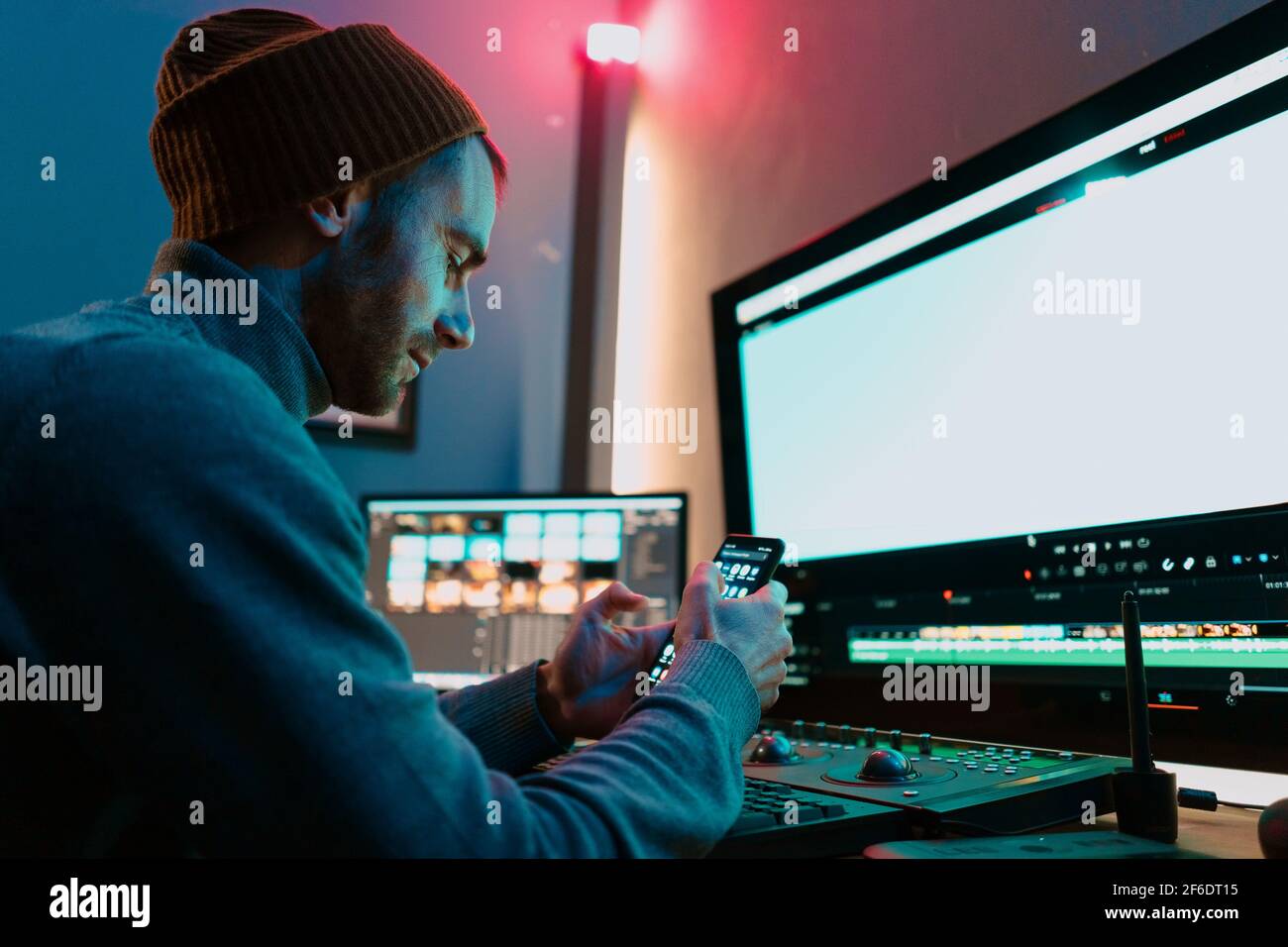 Attractive Male Video Editor Works with Footage or Video on His Personal Computer and having a break communicating on his smartphone. He Works in Creative Office Studio or home. Neon lights Stock Photo