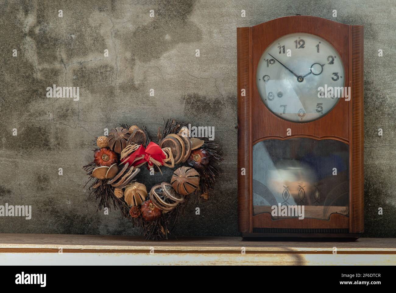 Heart shape wreath made from dried fruits placed with vintage wood pendulum clock which is leaning against the cement wall. Concept of "There's always Stock Photo