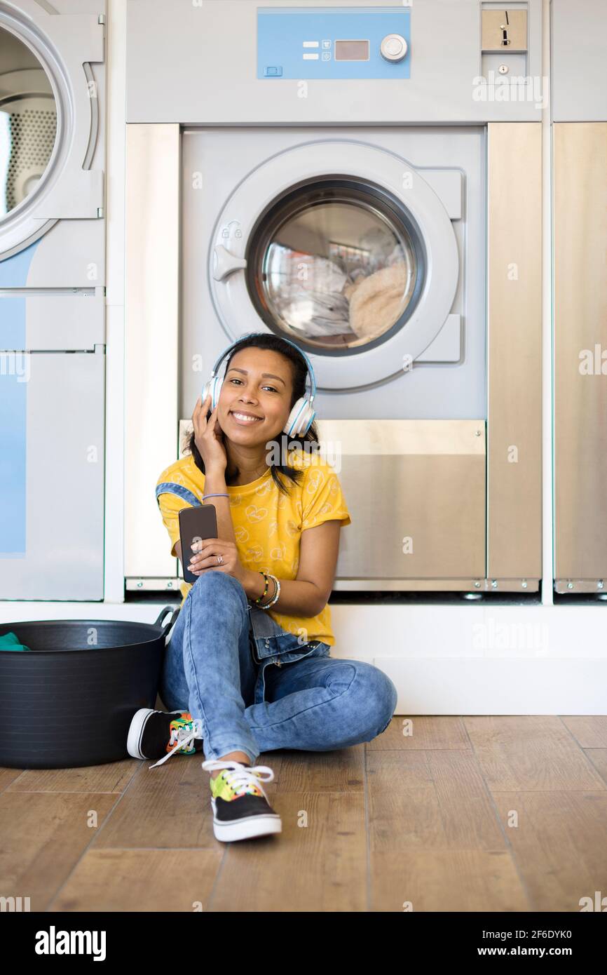 Young hispanic woman enjoying music waiting for the clothes to be washed sitting on the floor at the self-service laundry. Space for text. Stock Photo