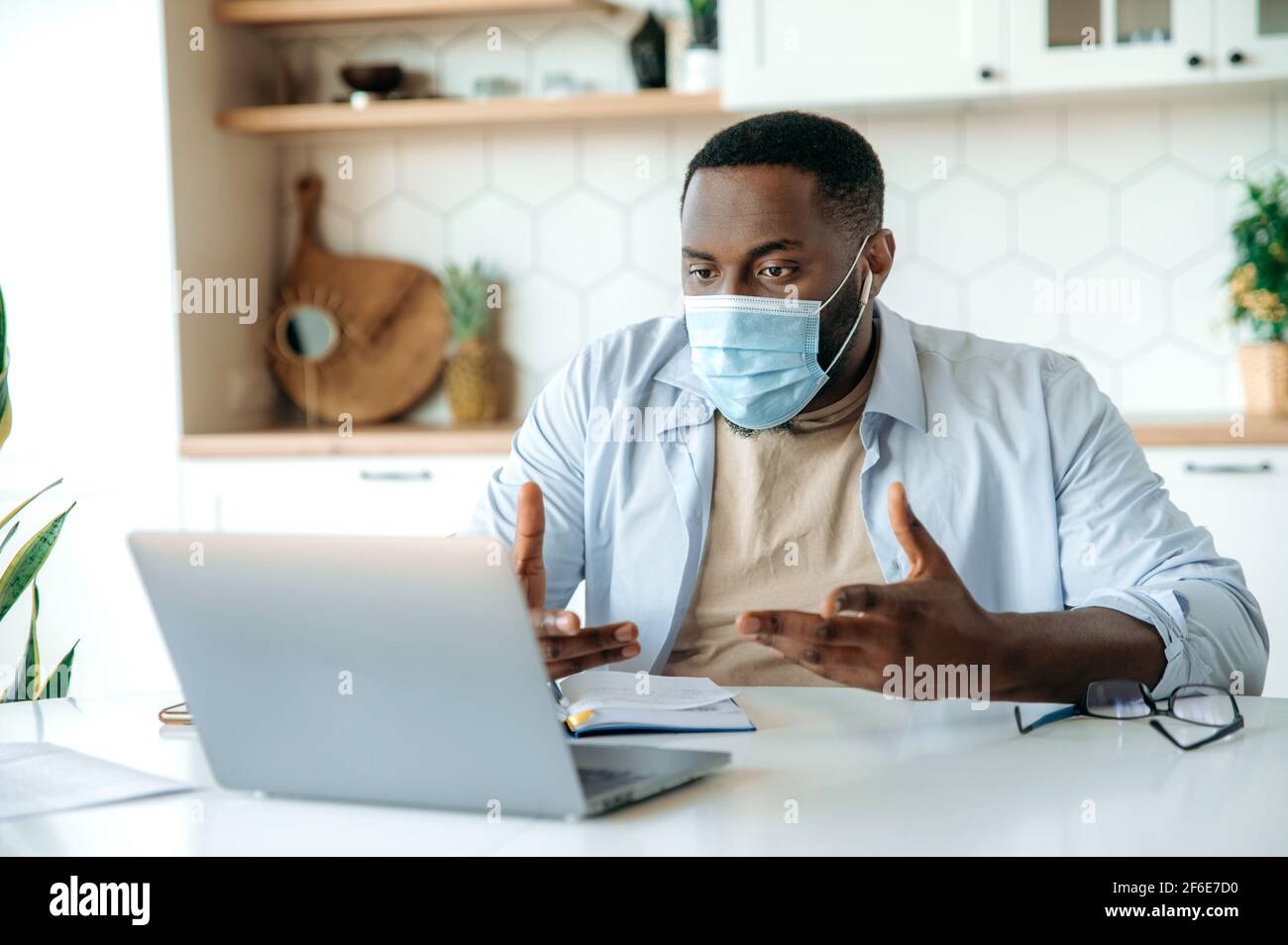 Work from home during quarantine. African American male freelancer or manager, wearing protective medical mask, working remotely, chatting with colleagues or clients via video conference by laptop Stock Photo