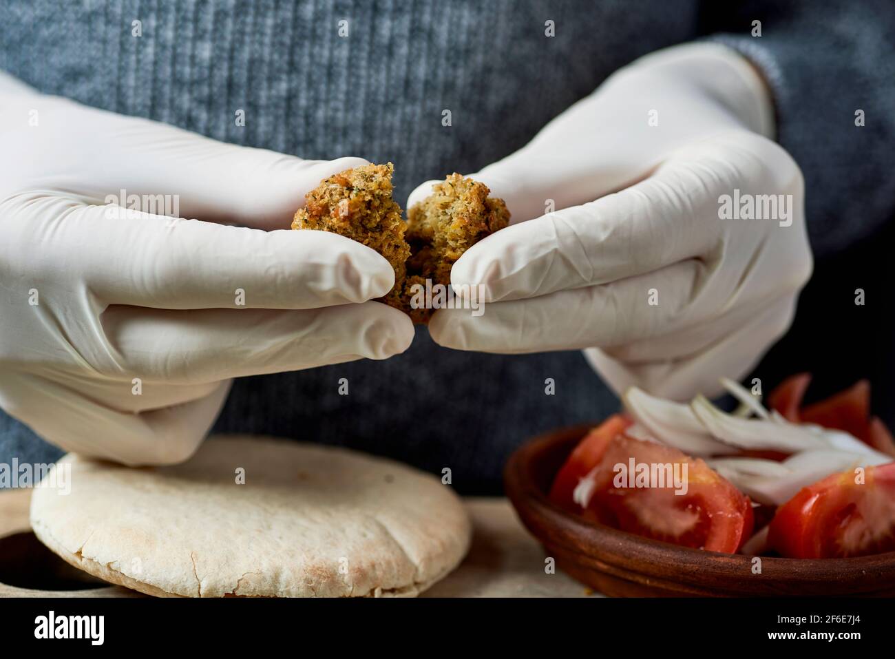 closeup of a young man, wearing latex gloves, about to prepare a falafel sandwich in a pita bread, with some chopped tomato and onion Stock Photo