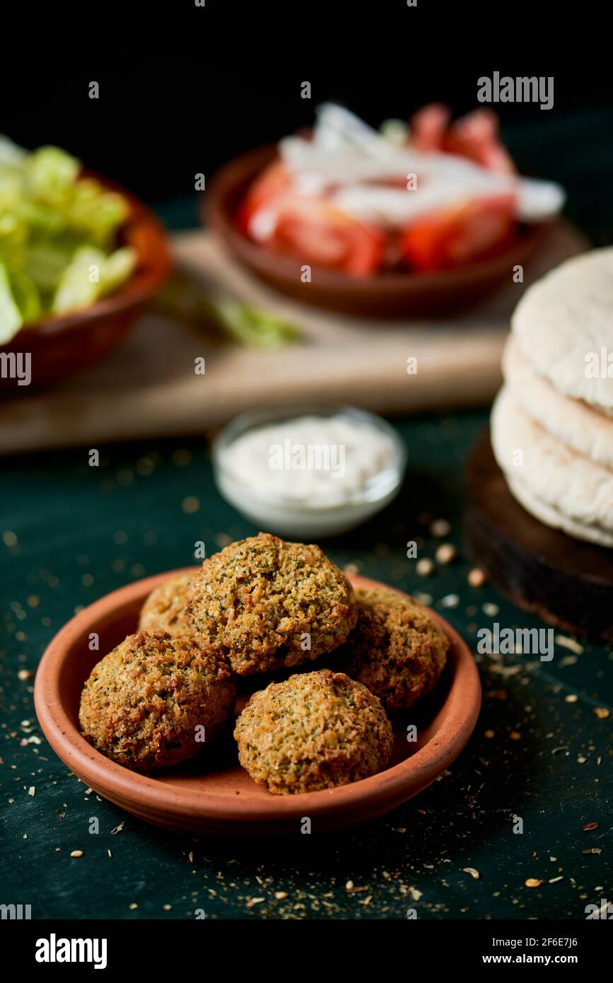 closeup of an earthenware bowl with some falafel, some chopped lettuce, onion and tomato in some plates and a pile of pita breads, on a rustic green w Stock Photo