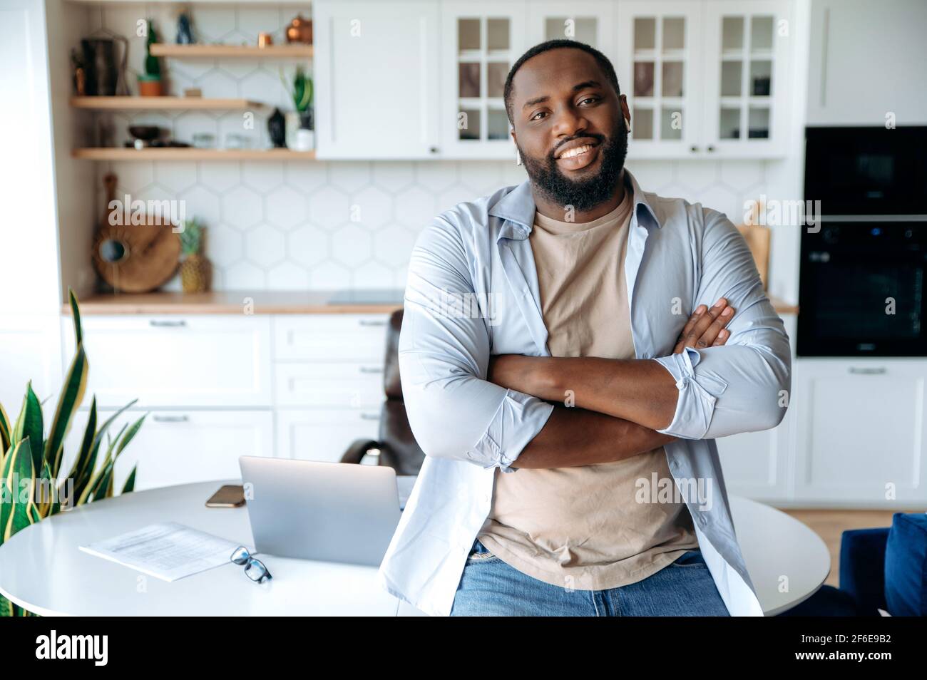 Friendly handsome confident stylish bearded African American male freelancer, standing near the workplace at home, crossing his arms, looking at the camera smiling friendly. Work from home concept Stock Photo