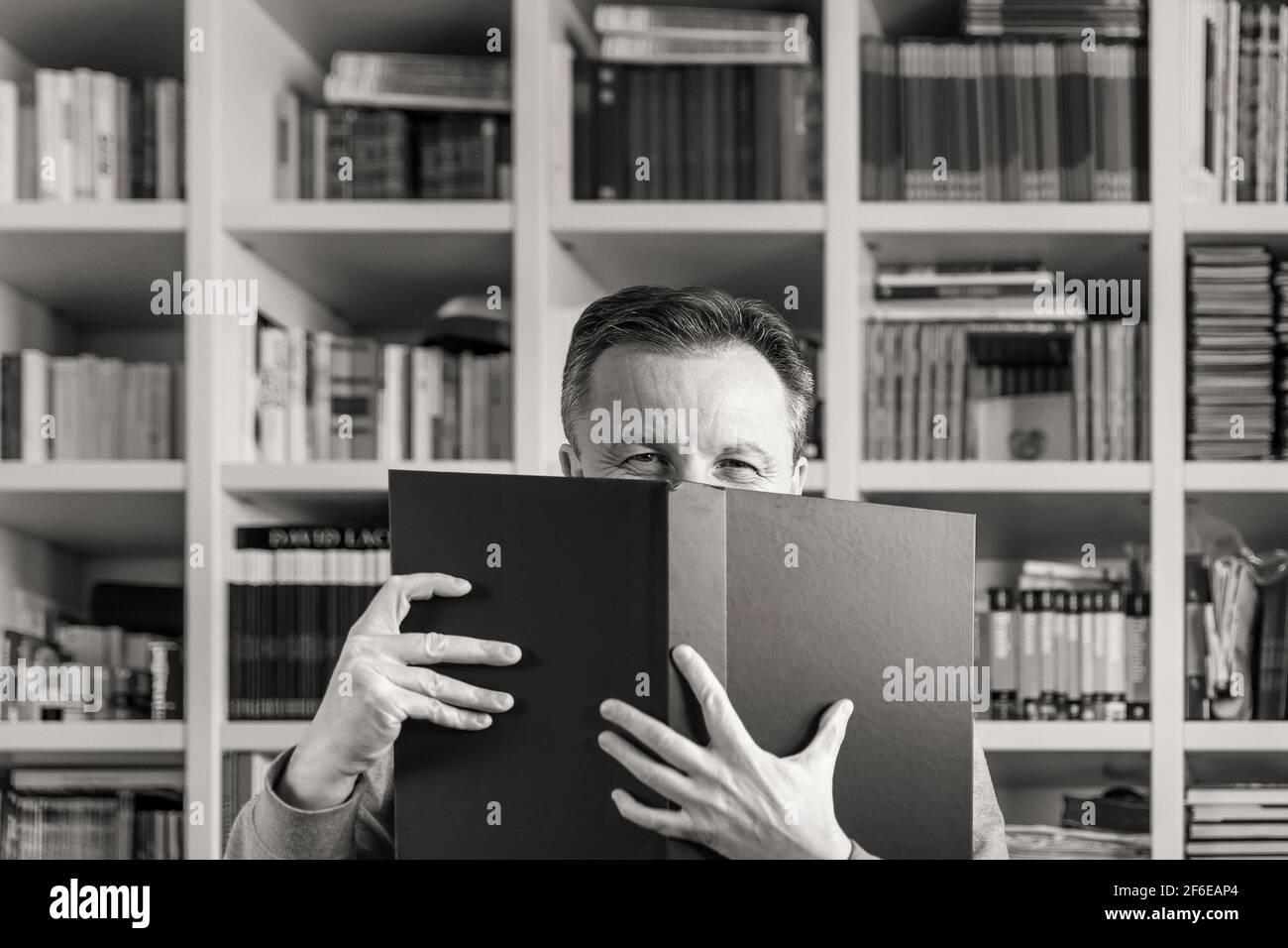 Black and white portrait of a man who has his face almost completely covered by a large book with a square bookcase in the background Stock Photo