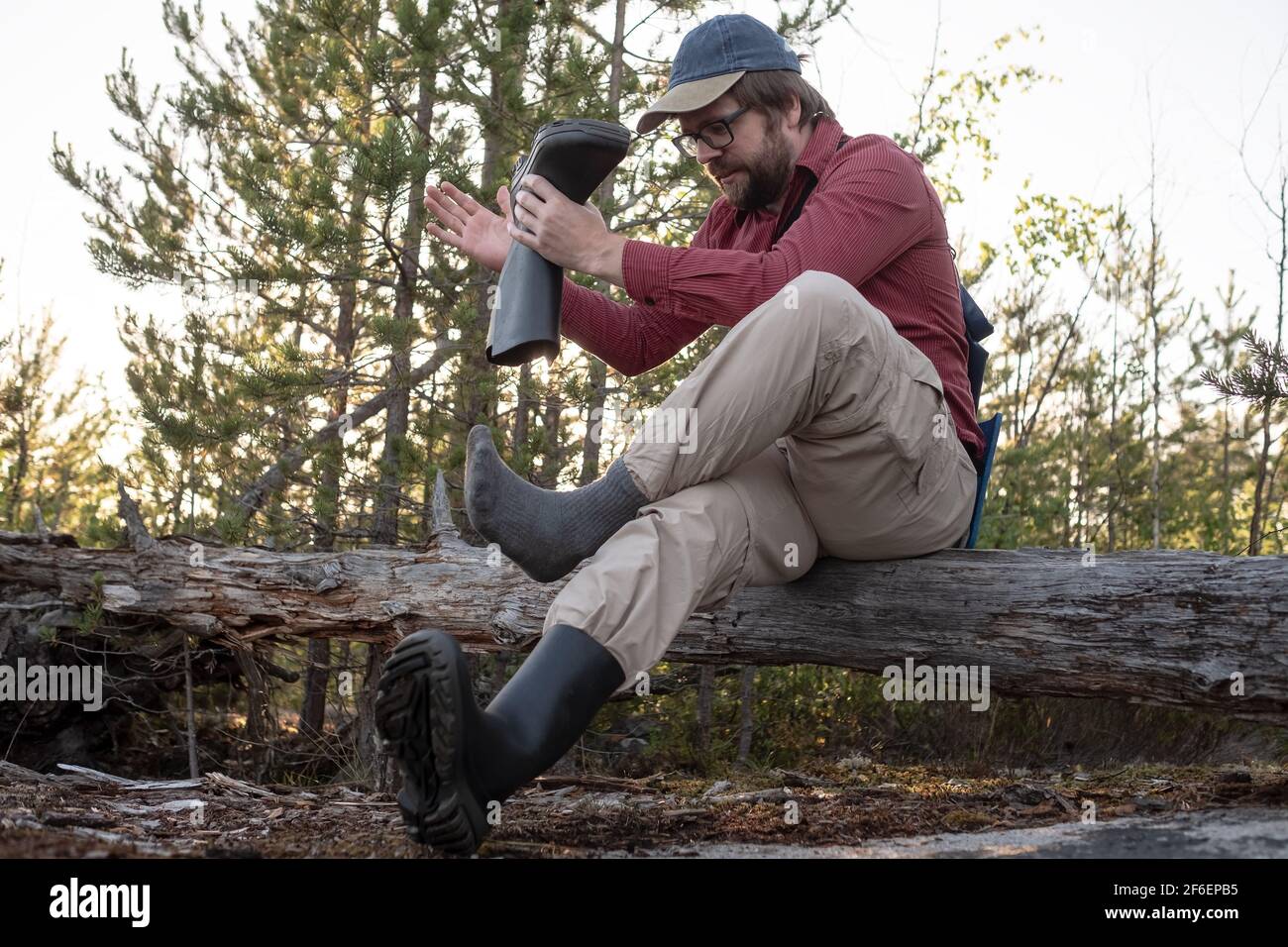 Man in hiking clothes sat on an old log in the woods, took off his rubber boots and shakes out trash. Active lifestyle concept. Stock Photo