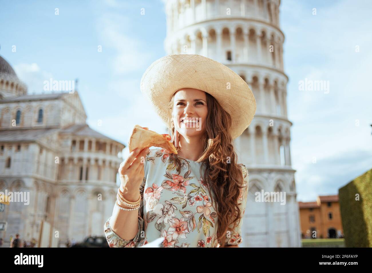 smiling stylish traveller woman in floral dress with pizza and hat in piazza dei miracoli in Pisa, Italy. Stock Photo