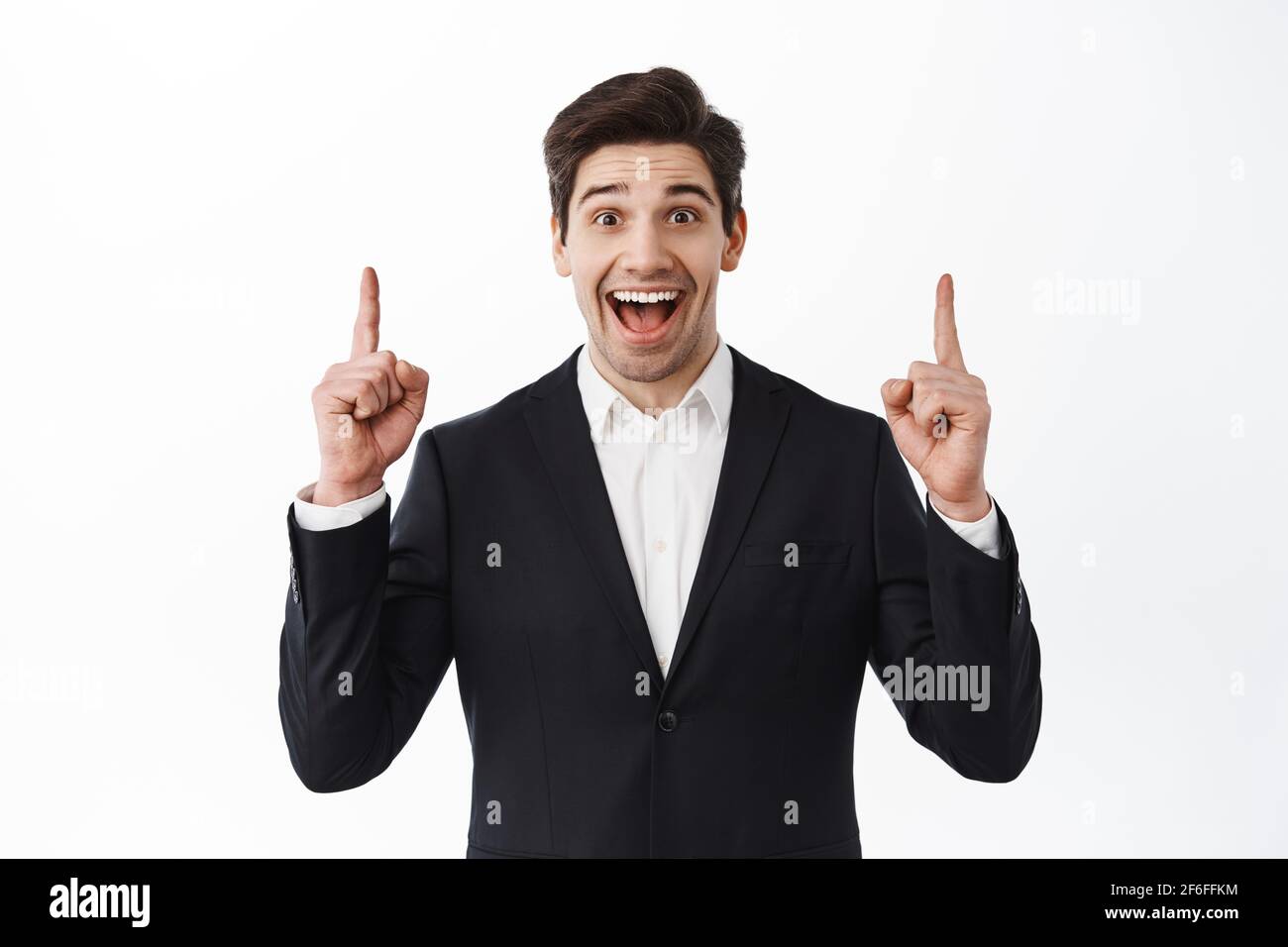 Cheerful male estate agent, broker in suit pointing fingers up, showing amazing offer, best promo sale, smiling amused, standing over white background Stock Photo