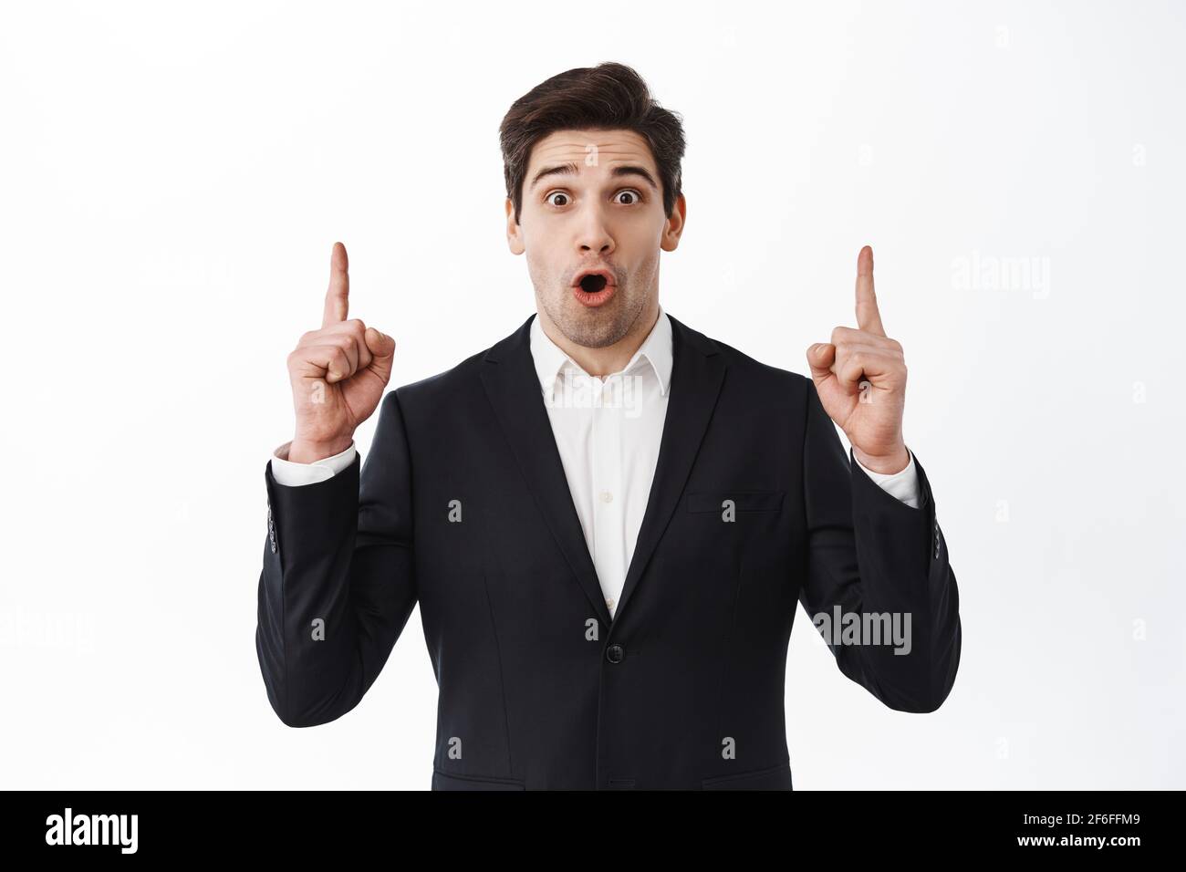 Impressed businessman, entrepreneur in black suit gasping, say wow and point up at copyspace, showing top advertisement, white background Stock Photo