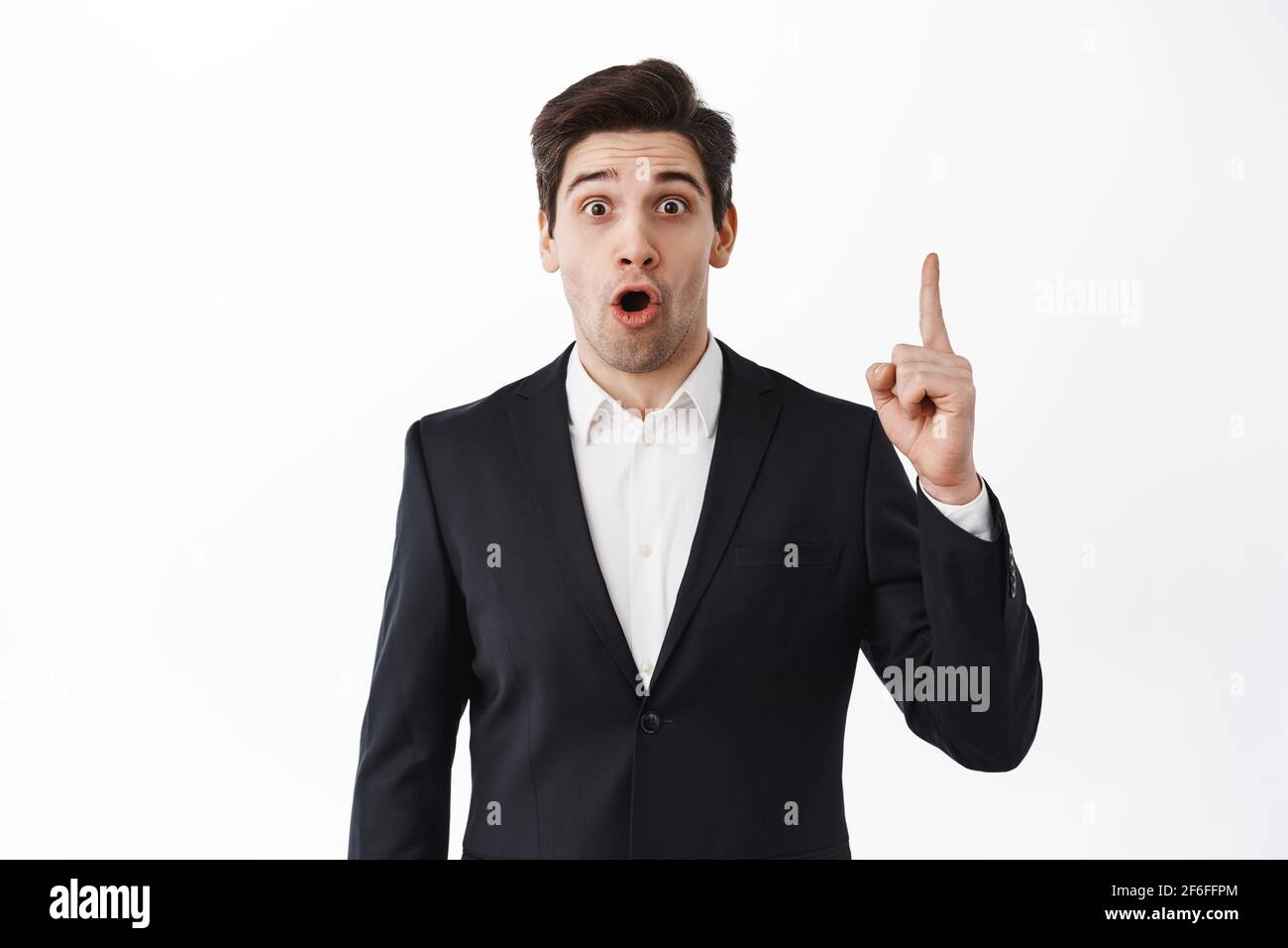 Surprised businessman pointing finger up and gasping amazed, say wow, stare excited at camera, standing in black suit against white studio background Stock Photo