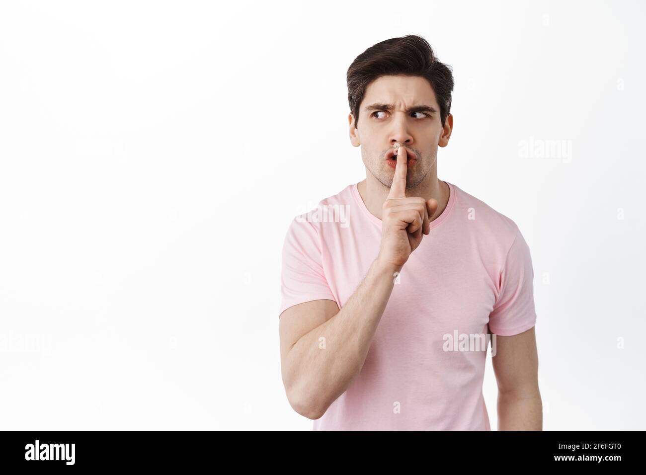Angry suspicious man looks aside and shushing, tell to shut up, be quiet, need silence, show taboo gesture, standing over white background Stock Photo
