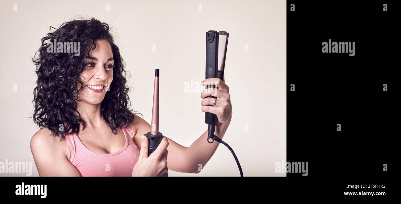 Smiling young curly-haired woman in profile looks at her hair curler and hair straightener. care and beauty concept. copy space in black Stock Photo