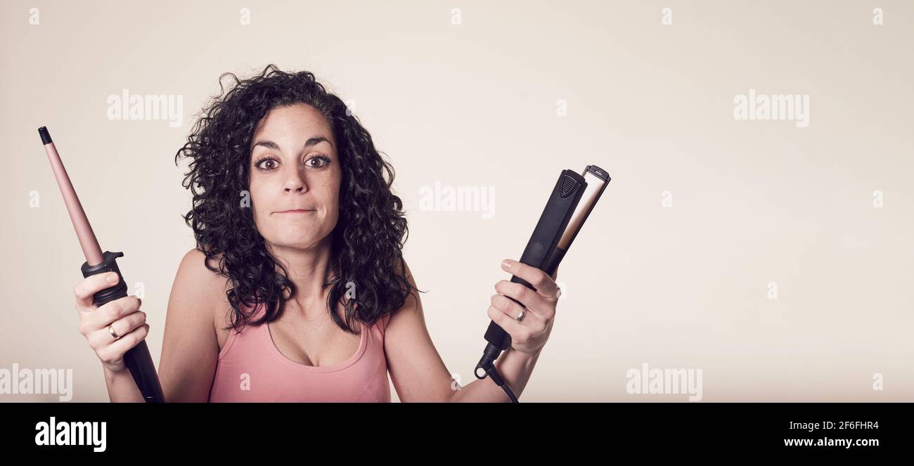 Young smiling curly-haired woman can't decide between using her curling iron or her hair straightener. doubt gesture. care and beauty concept. Stock Photo