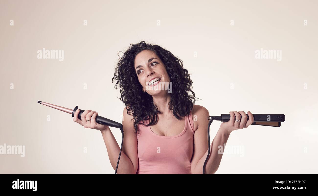 Young smiling curly-haired woman can't decide between using her curling iron or her hair straightener. doubt gesture. care and beauty concept. Stock Photo