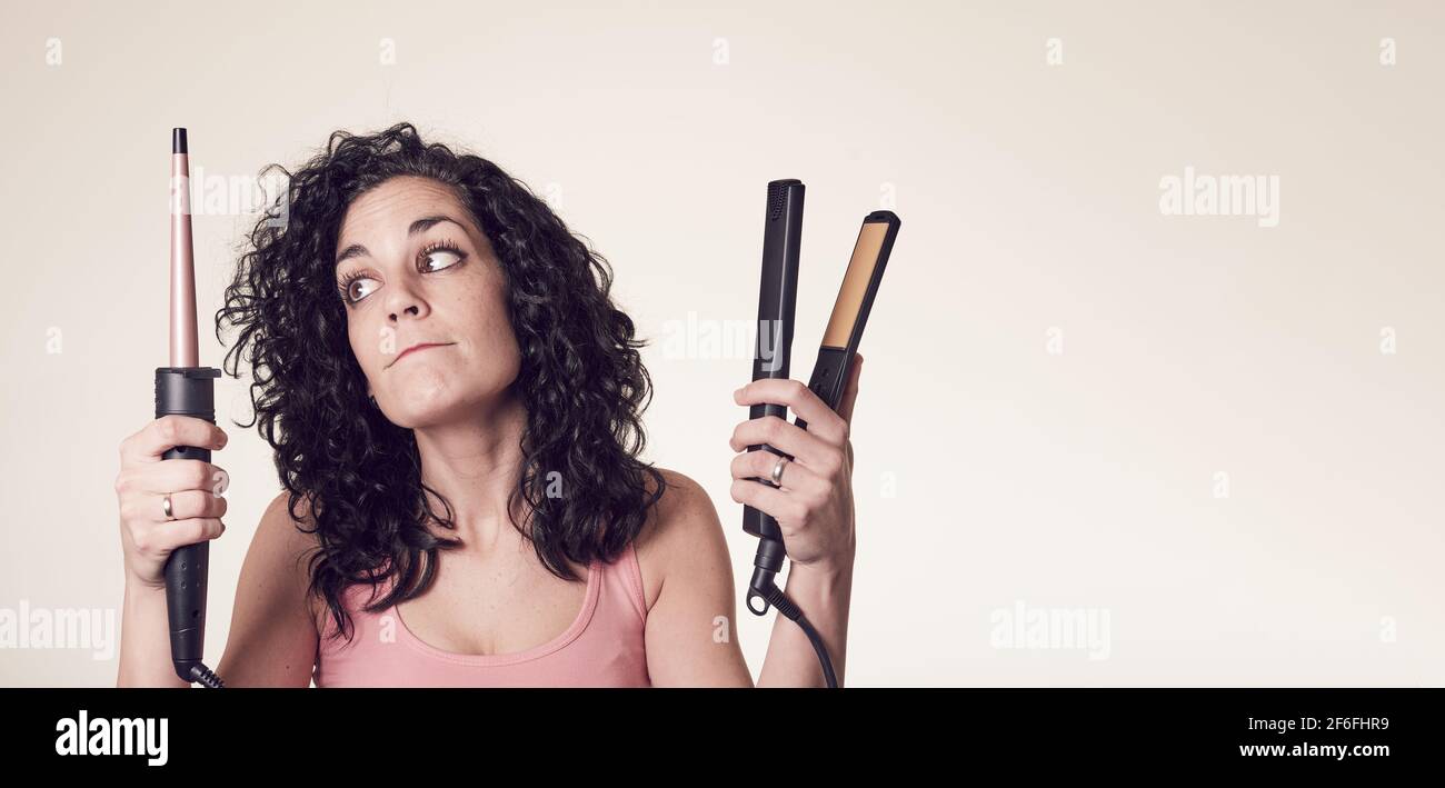 Young smiling curly-haired woman can't decide between using her curling iron or her hair straightener. doubt gesture. care and beauty concept.Young sm Stock Photo