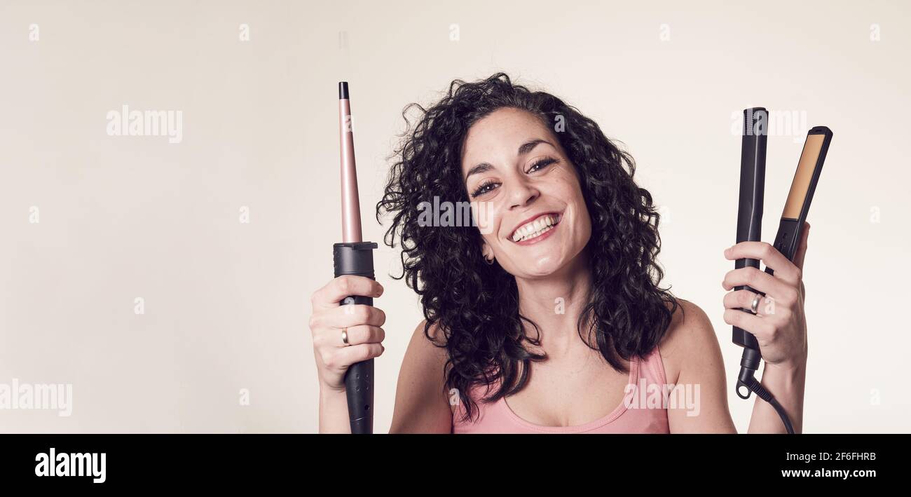Smiling curly-haired young man can't decide between using his curling iron or his hair straightener. care and beauty concept copy space. Stock Photo