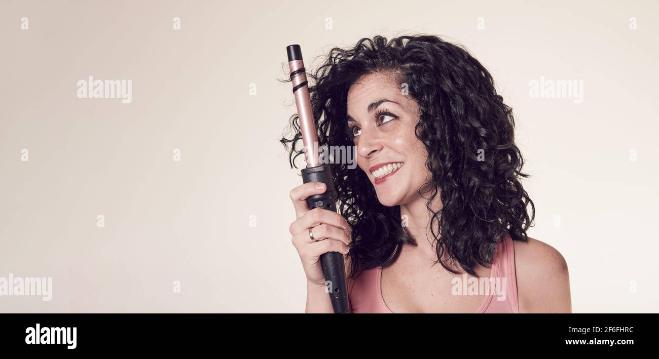 Young smiling woman with black curly hair uses tweezers or curling iron to get curls and waves in her hair. care and beauty concept. copy space. Stock Photo