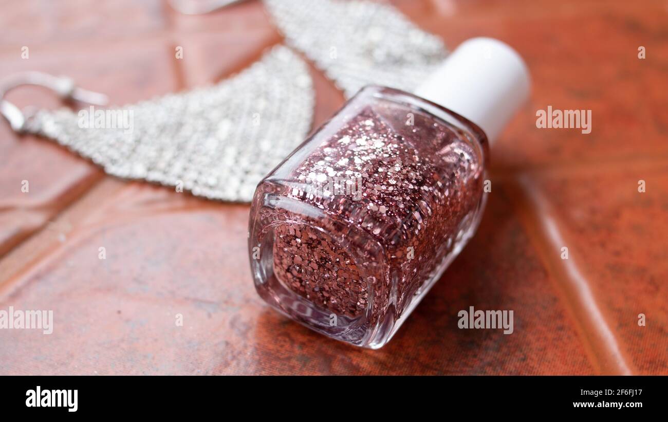 Rose gold sparkle nail polish, soft-focus, laying on terracotta tile next to a pair of sparkly diamond earrings, Ontario, Canada, 2021. Stock Photo