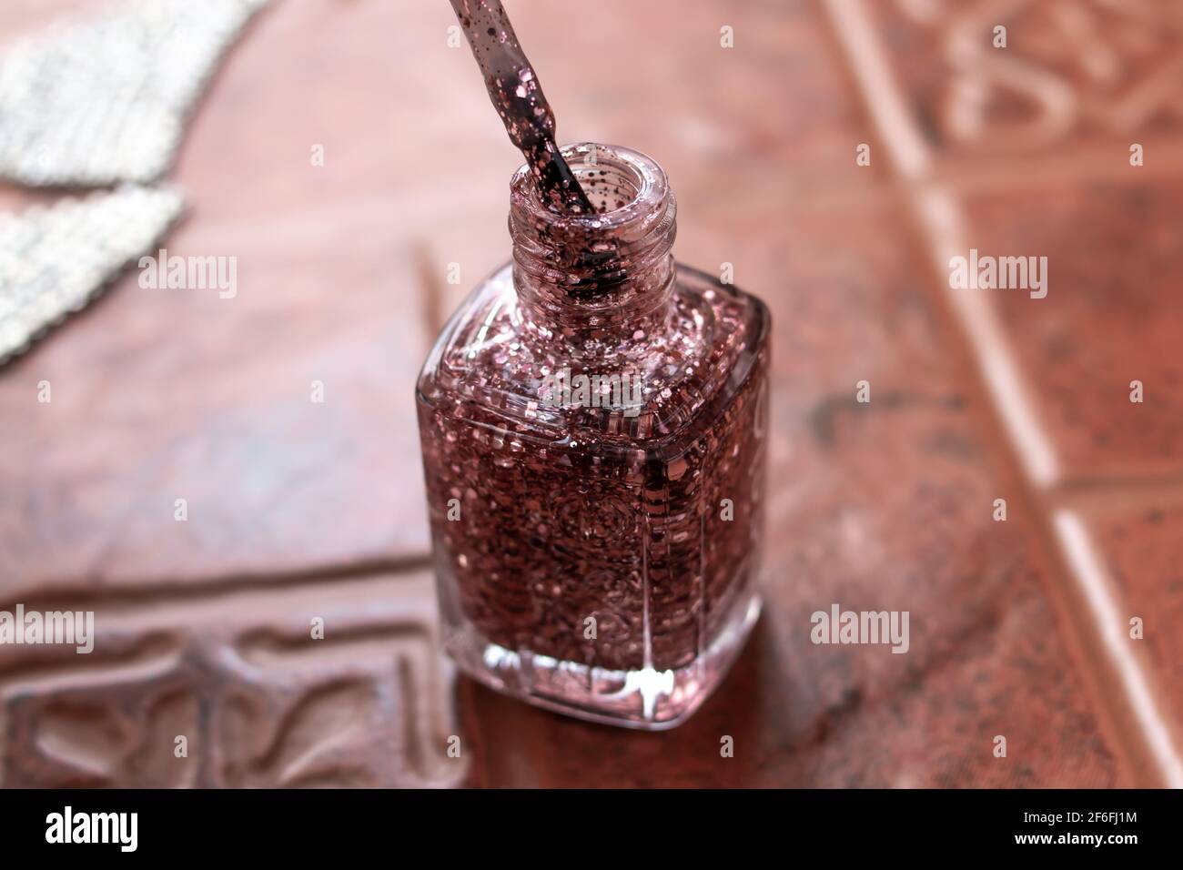 Rose gold sparkle nail polish, soft-focus in a clear glass bottle with the polish brush uncapped and leaving the bottle. Terracotta tile background. Stock Photo