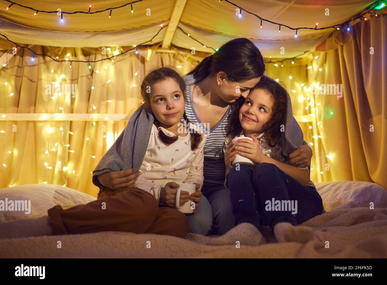 Mother hugs two daughters drinking tea or milk in kids tent before bedtime Stock Photo