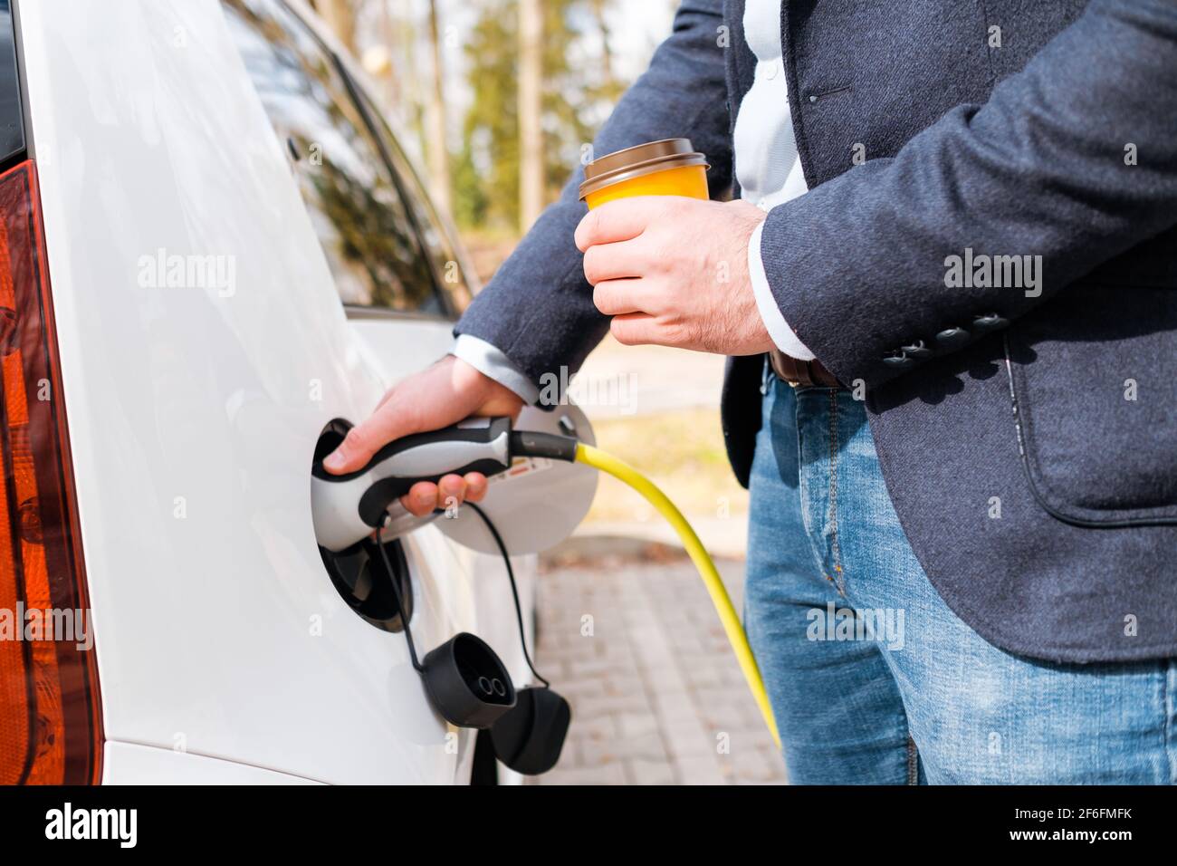 Close up man inserting power cable supply for charging electric car and holding cup of coffee.  Stock Photo