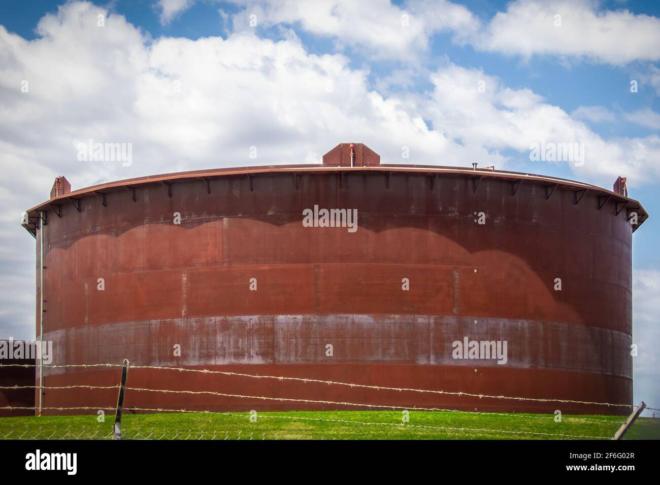 Giant rusty oil tank in tank farm in Cushing Oklahoma behind barbed wire fence with pretty blue sky background Stock Photo