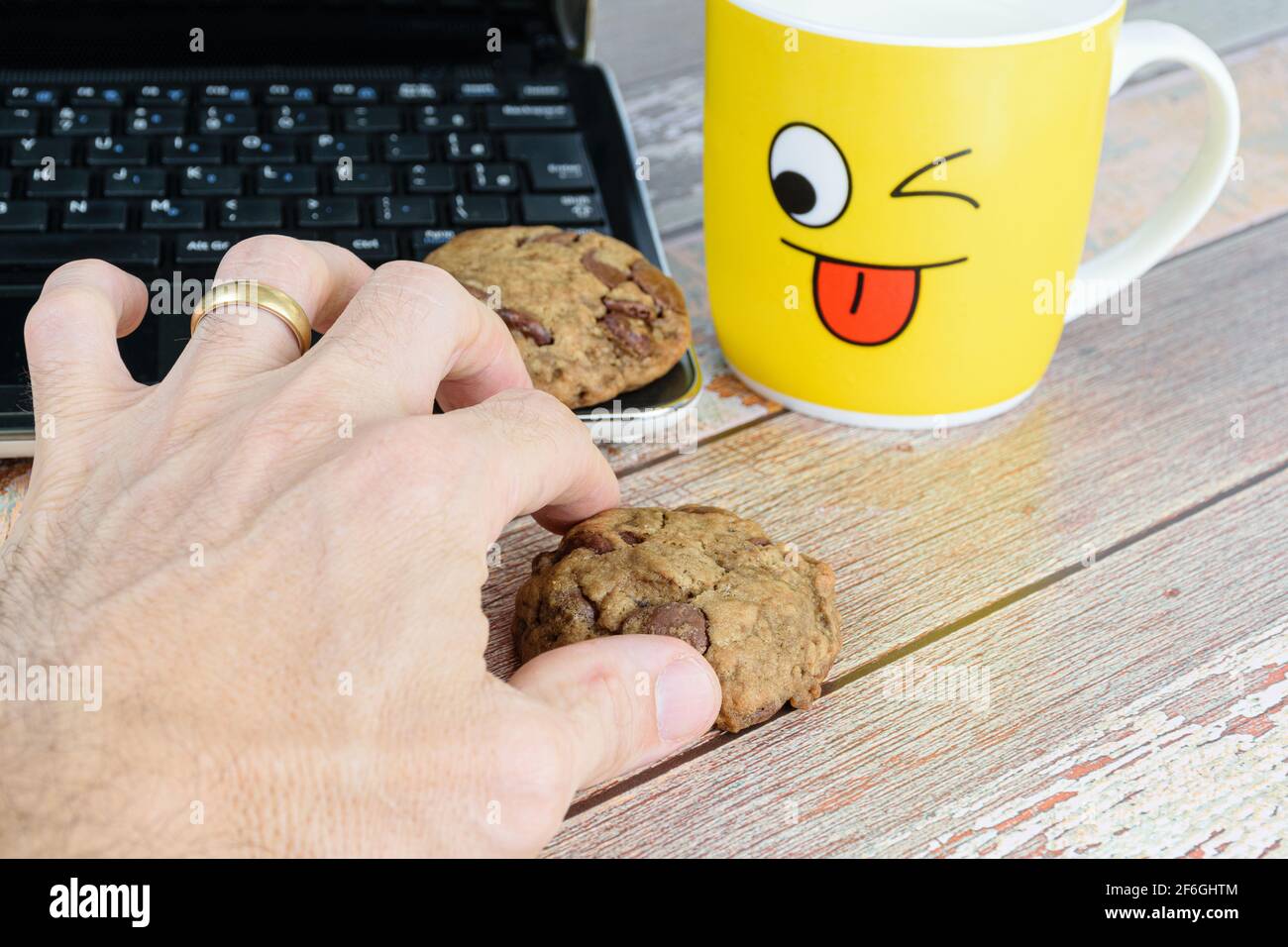 Male hand picking up a cookie with drops of chocolates. In the background, a yellow mug and a laptop. Pause for e-learning. Stock Photo
