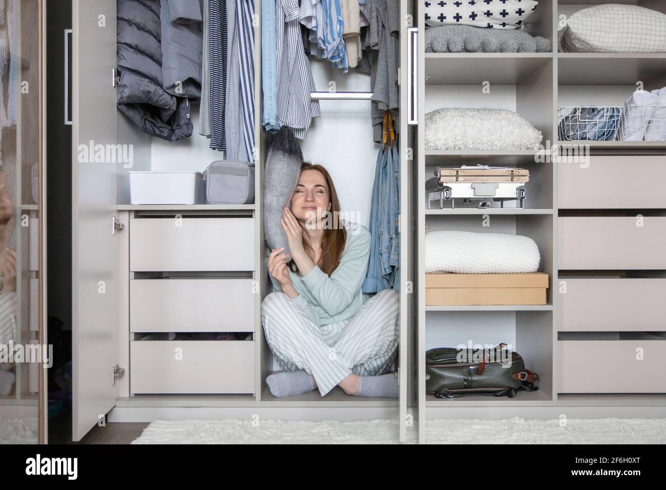 Smiling housewife in pajamas sleeping on pillow at wardrobe. Woman hiding in modern cupboard Stock Photo