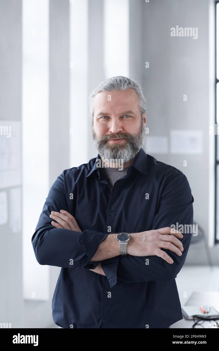 Portrait of mature bearded businessman smiling at camera while standing with his arms crossed at office Stock Photo