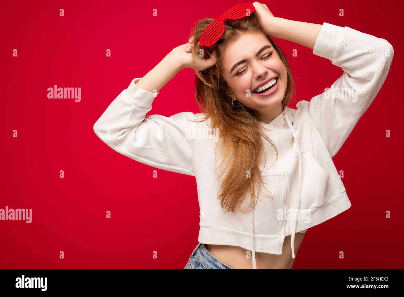 Shot of beautiful happy smiling young blonde woman isolated over red background wearing casual white hoodie and stylish sunglasses enjoying and having Stock Photo