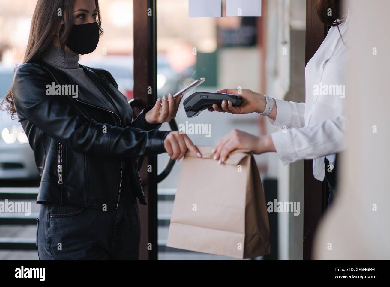 Young woman standing outdoors in mask and paying bill through smartphone using NFC technology in a cafe. Female customer paying using contactless Stock Photo