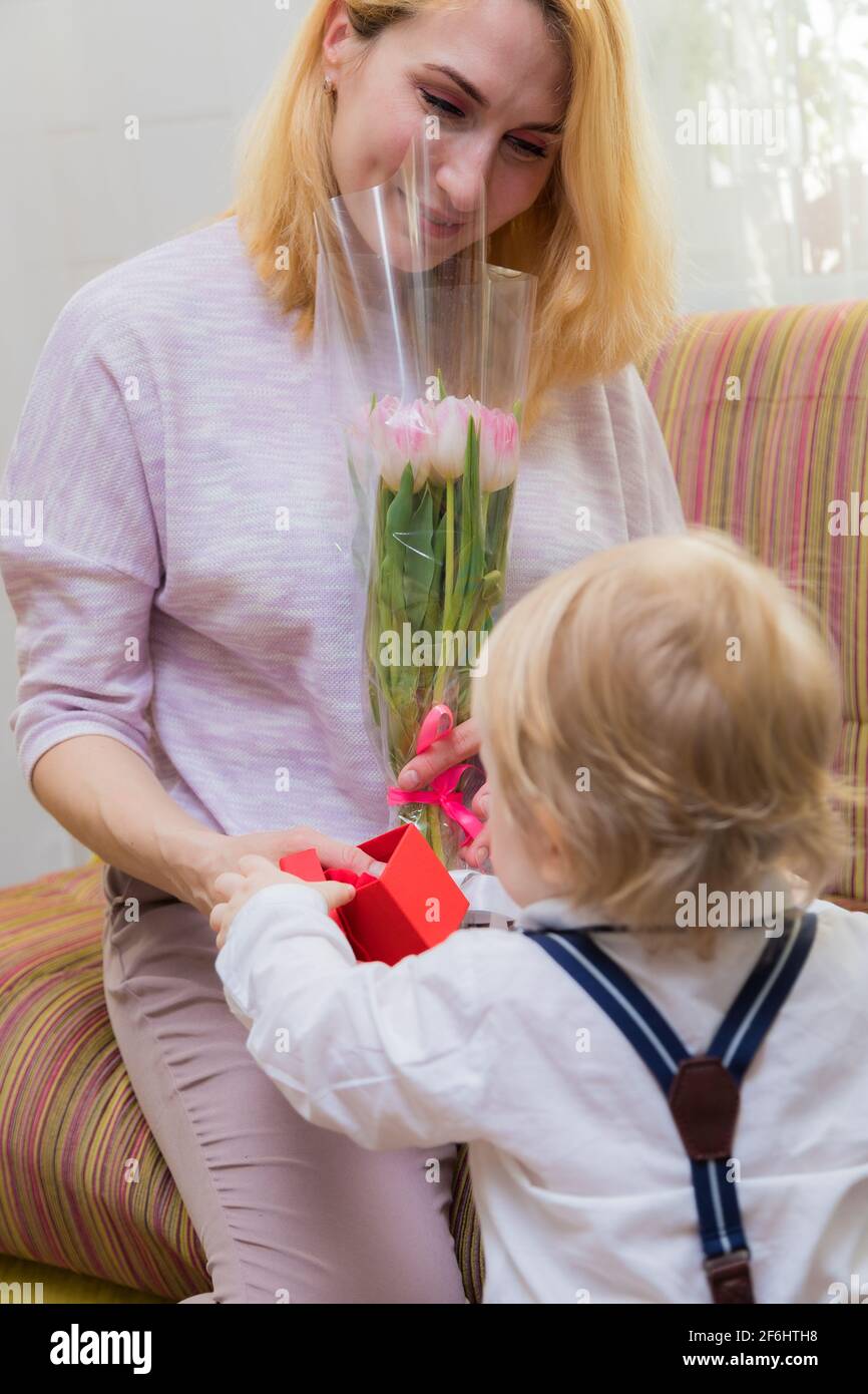 The kid congratulates his mother on the holiday, gives her flowers and a gift. Boy in white shirt and trousers with suspenders Stock Photo