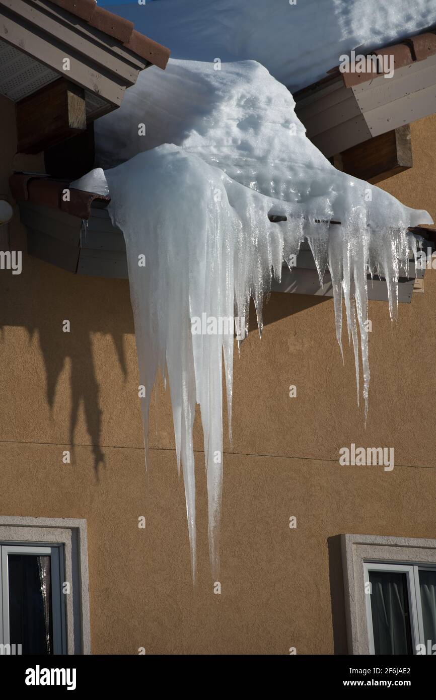 very large frozen icicle hanging from exterior home eaves of roof casting shadow on exterior  home concrete wall snow load visible freeze thaw climate Stock Photo