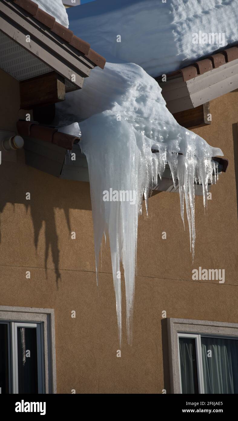 very large frozen icicle hanging from exterior home eaves of roof casting shadow on exterior  home concrete wall snow load visible freeze thaw climate Stock Photo