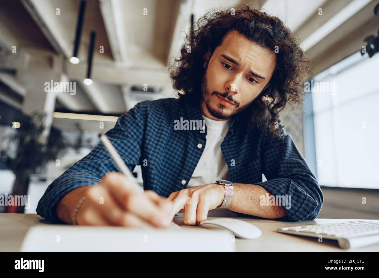 Handsome curly guy is making notes at the working table in open space office Stock Photo