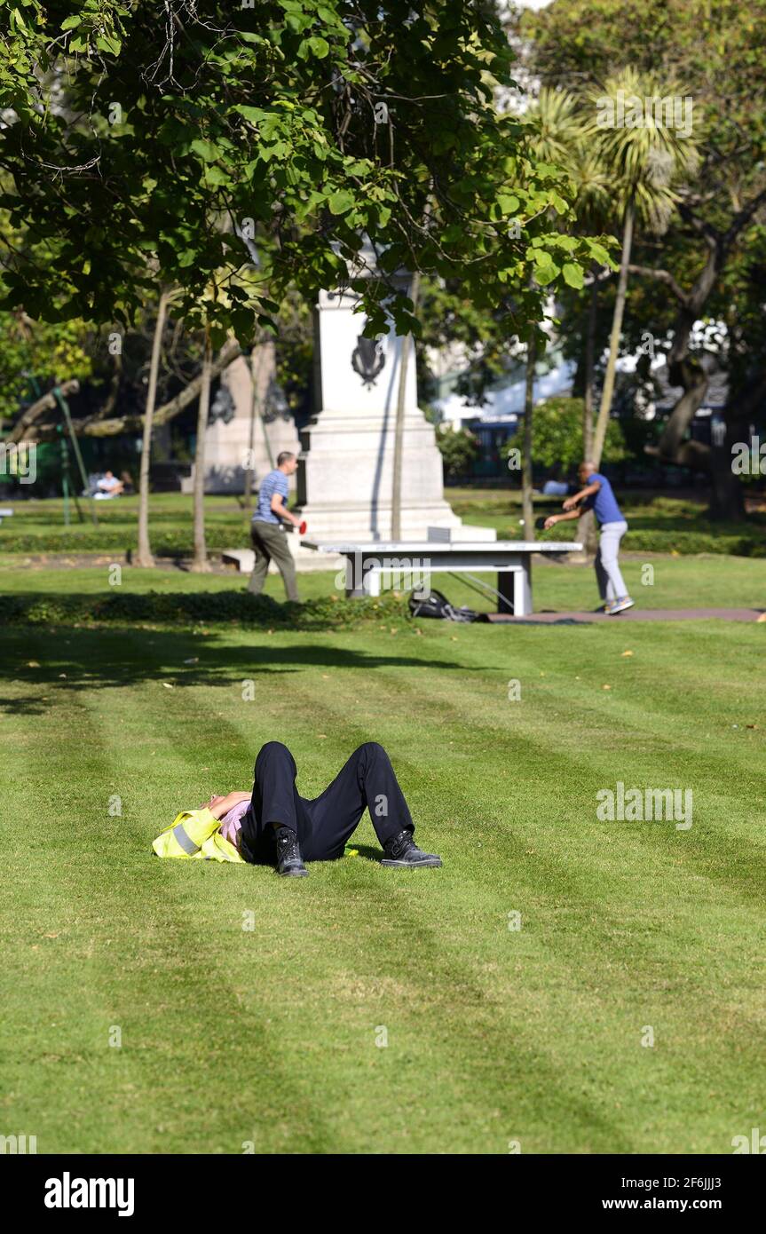 London, England, UK. Man in a hi-vis jacket relaxing on the grass in Victoria Embankment Gardens as two men play outdoor table tennis Stock Photo